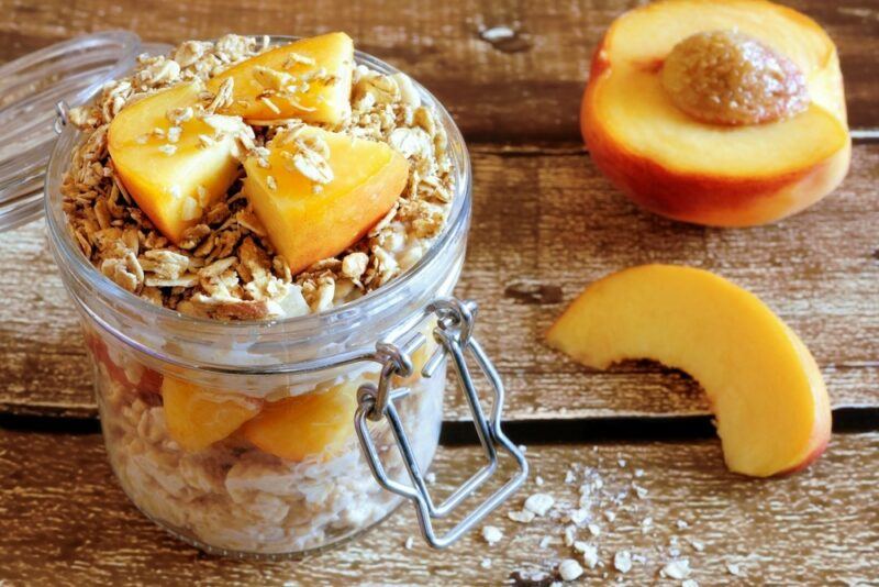 A wooden table with a jar of overnight oats and peaches, next to a fresh peach