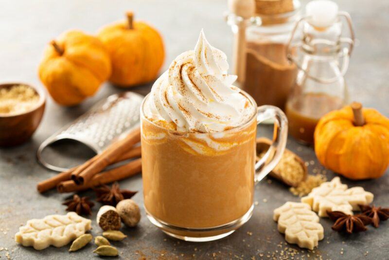 A glass mug containing a pumpkin spice latte with two fresh pumpkins in the background, cookies and other ingredients on the table