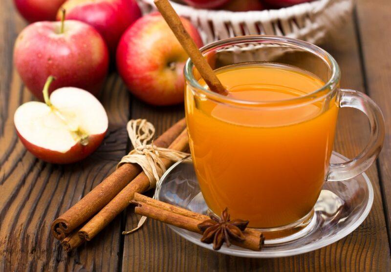 Una taza de vidrio de sidra de manzana con palitos de canela, junto a algunas manzanas frescas