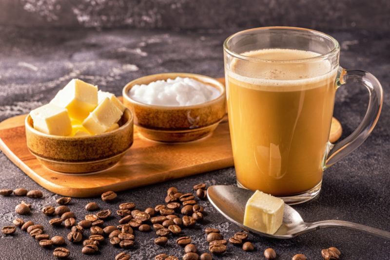 A wooden board with a small bowl of butter and one of coconut oil, next to a spoon of butter and a mug of Bulletproof coffee