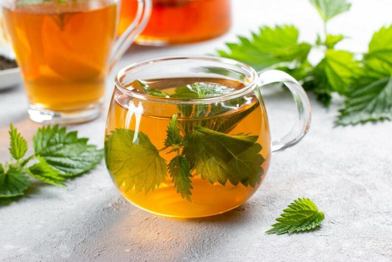 A glass of fresh herbal tea with the herbs still in it, with another couple in the background and some herbs scattered on the table