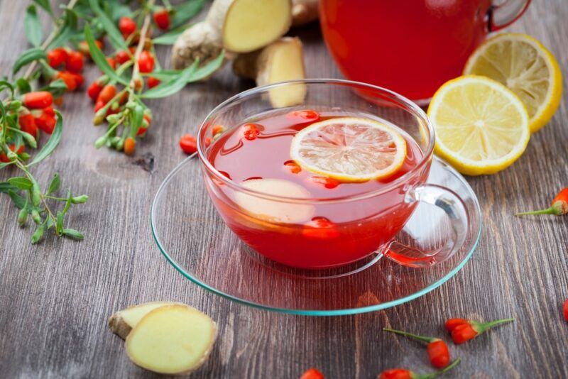 A glass mug filled with goji berry tea, with fresh goji berries and lemons in the background