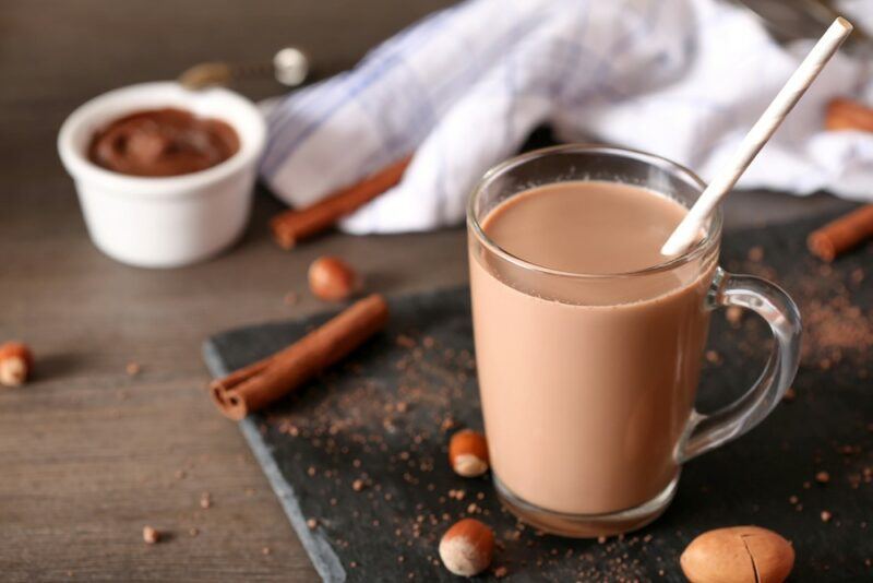 A glass of homemade chocolate milk, made using cinnamon and hazelnuts. There is a small tub of chocolate sauce in the background