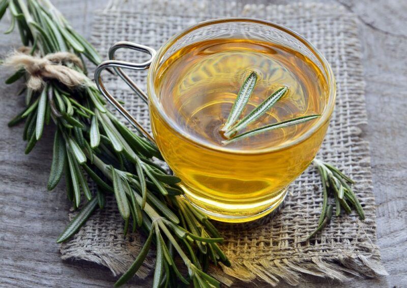 A wooden table with a piece of cloth, with a glass mug of rosemary tea, next to a sprig of rosemary