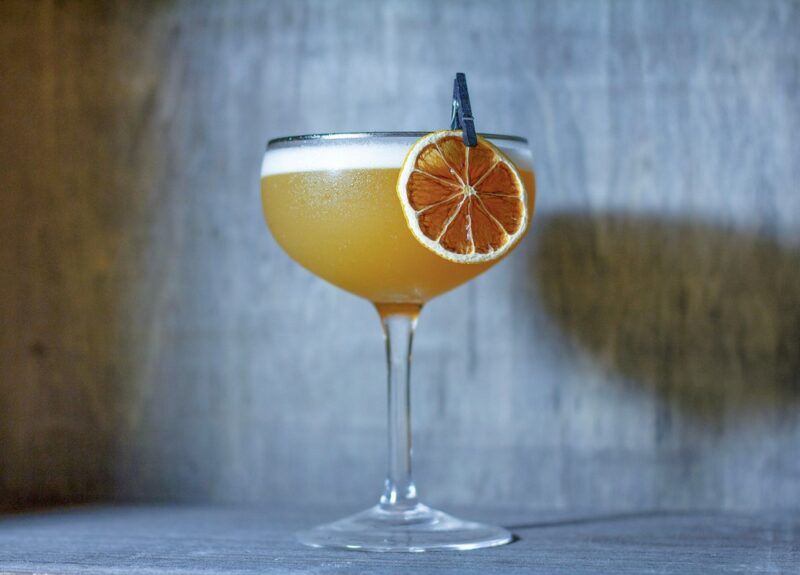 A Kyoto sour cocktail against a grey wall, using a slice of dried orange or blood orange as a garnish