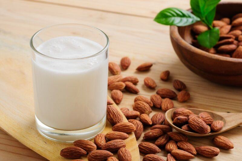 A glass of almond milk on a wooden board next to almonds and a bowl of almonds