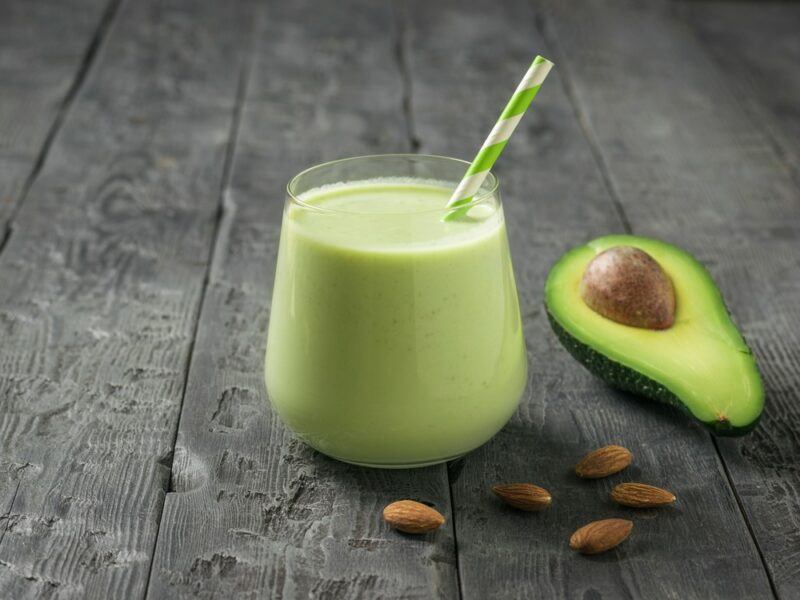 A wooden table with a glass of avocado smoothie, next to an avocado and a handful of almonds