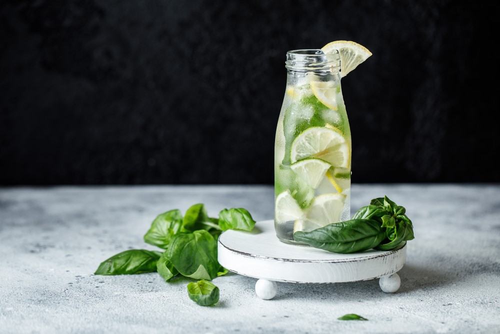 A glass bottle of basil water with lemon and limes, next to fresh basil against a black background