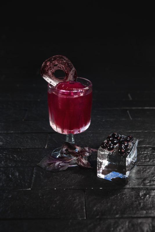 A glass of beet cocktail garnished with beetroot next to a glass of berries against a dark background