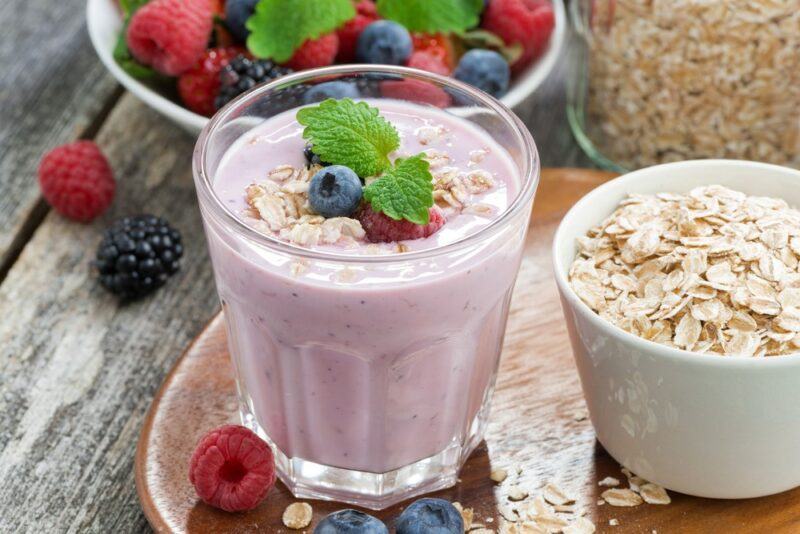 A glass of a berry smoothie, in front of a bowl of fruit and next to a bowl of oats