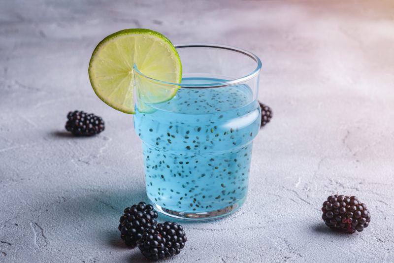 A glass containing a blue cocktail with chia seeds that has been garnished with lime. There are a few berries on the table