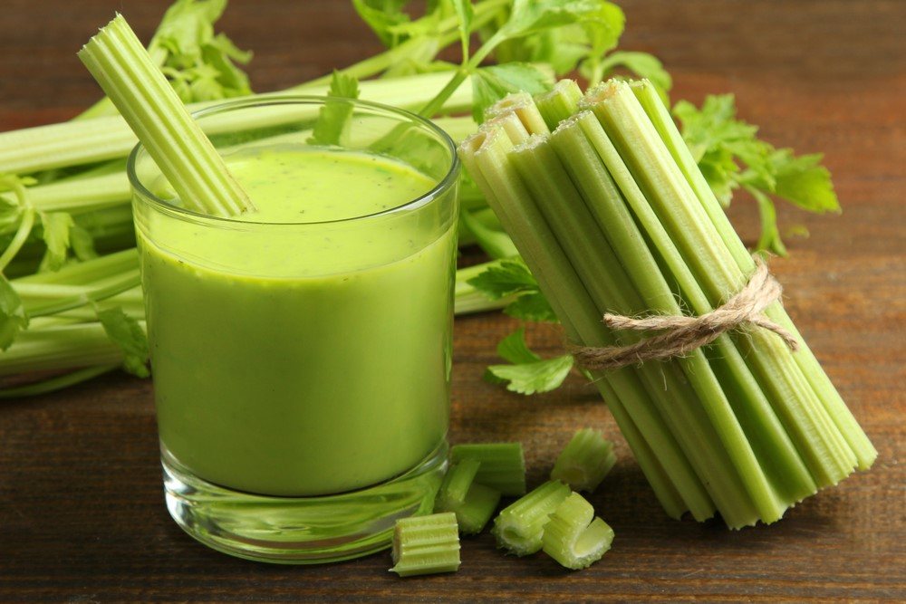 A glass of celery juice with celery sticking out of it, next to a bunch of celery and more in the background