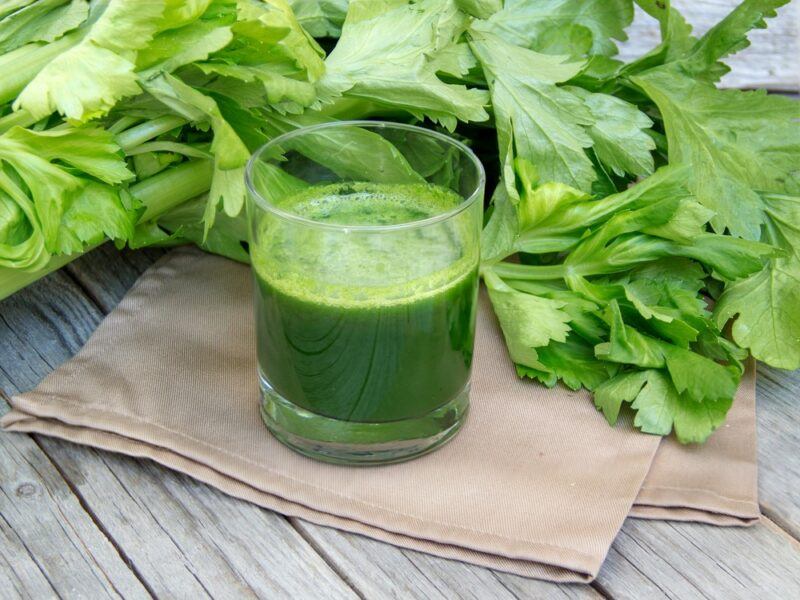 A wooden table with a large sprig of celery, a cloth, and a glass of celery juice