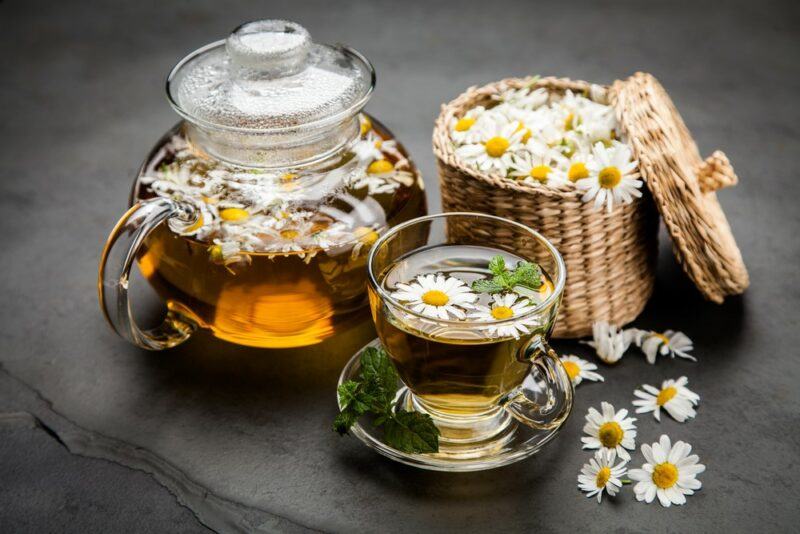 A black table with a pot of chamomile tea, a basket of the flowers, and a glass mug of the tea