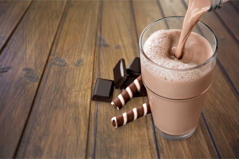 A wooden table or floor with a glass of chocolate milk being poured from a bottle, next to some straw and chocolate