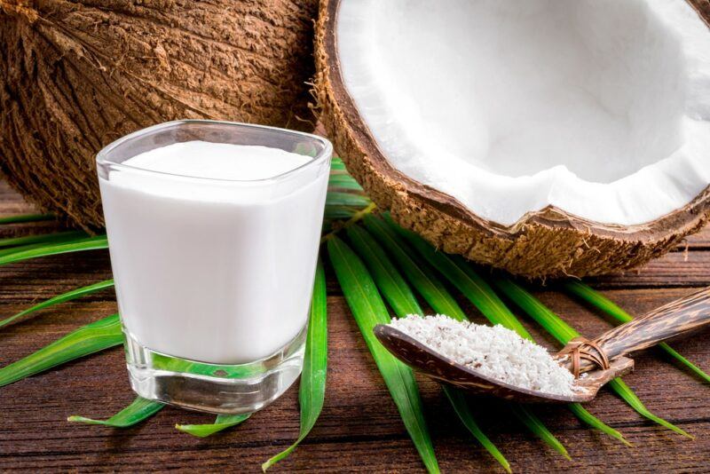 A small glass of coconut milk next to a spoon, in front of one whole coconut and one coconut half