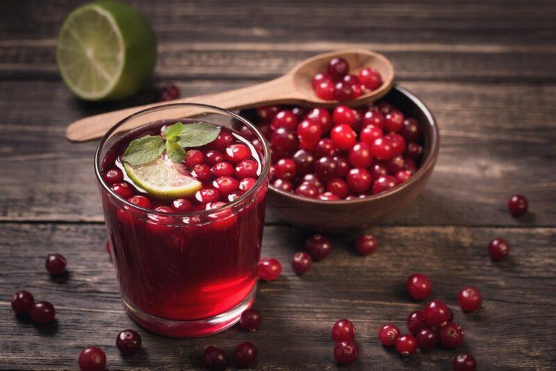 A dish of cranberries with a wooden spoon on top, behind a glass of cranberry juice and cranberries