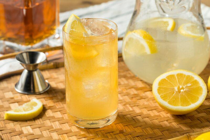 A glass of dragonade, next to a pitcher of lemonade, a cocktail jigger, and some lemons, with whiskey in the background