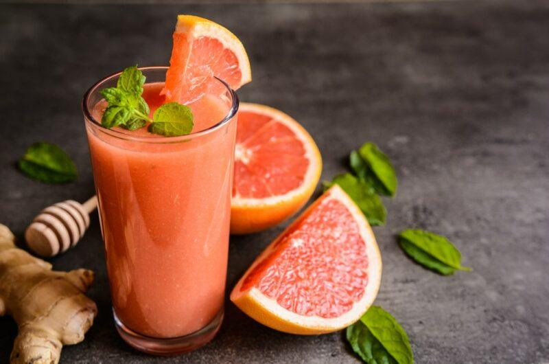 A black table with a glass of grapefruit juice, next to some grapefruit wedges, ginger, and a honey stick