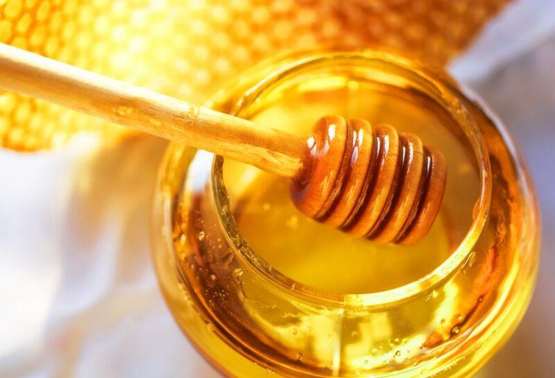 A glass jar of honey with a stirrer leaning on top, next to a gold colored piece of foil or paper