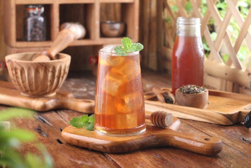 A wooden table with a glass of iced tea, with a pitcher of the tea in the background, plus a mortar and pestle, honey, and tea leaves