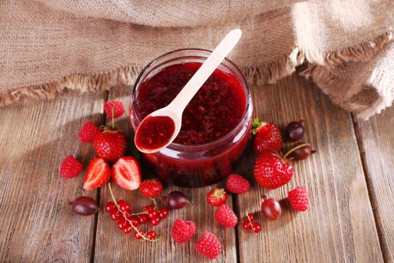 A wooden table with a jar of fresh jam, a spoon of jam, and plenty of fresh fruit