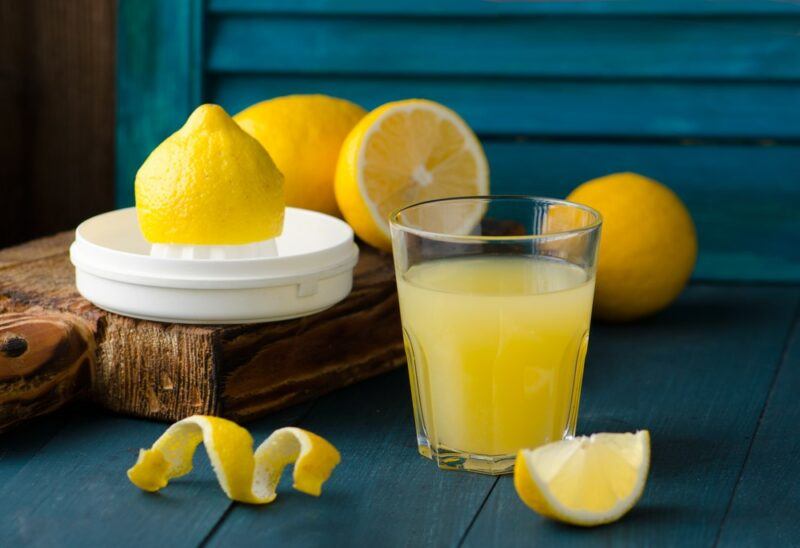 A glass of freshly squeezed lemon juice on a table, next to some lemons and lemon pieces, once that is still on a squeezing tool