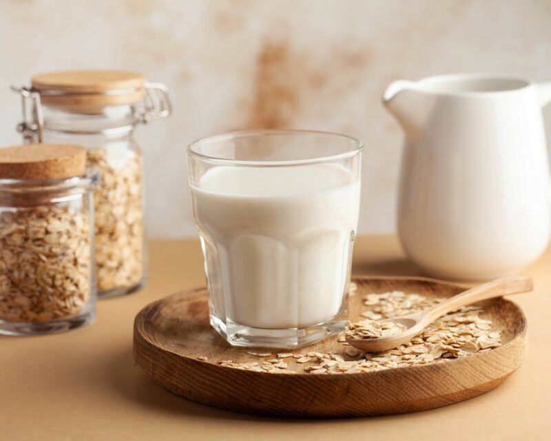 Two jars with lids containing oat milk, on a wooden tray with oats, a spoon, and a glass of oat milk, with a white jug in the background