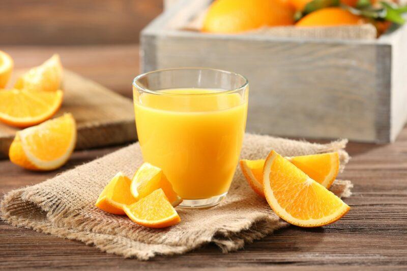 A glass of orange juice, with a selection of orange wedges on the table. There's a crate of oranges in the background