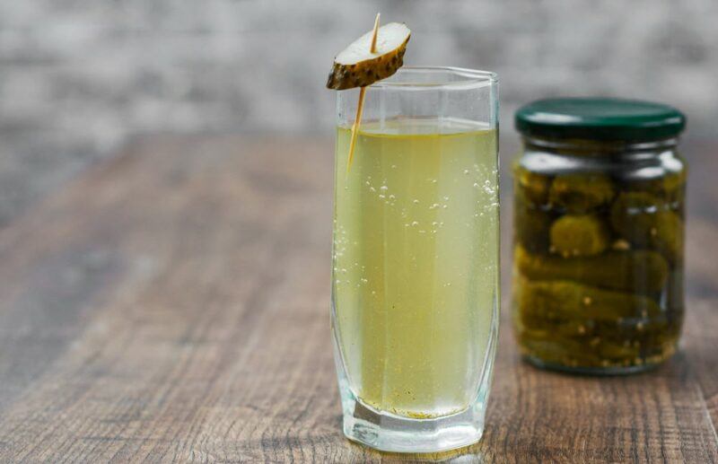 A wooden table with a glass of pickle juice garnished with a pickle slice, plus a sealed jar of pickles