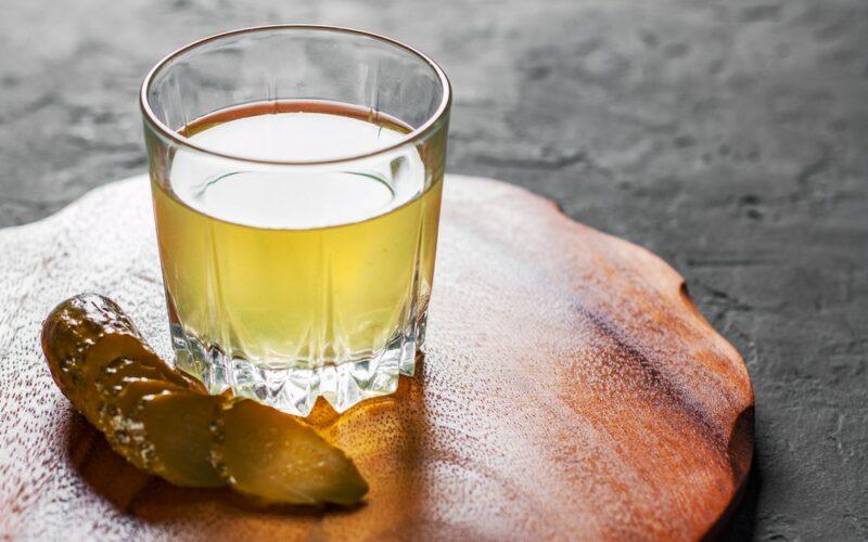 A wooden plan with a glass of pickle juice next to a pickle