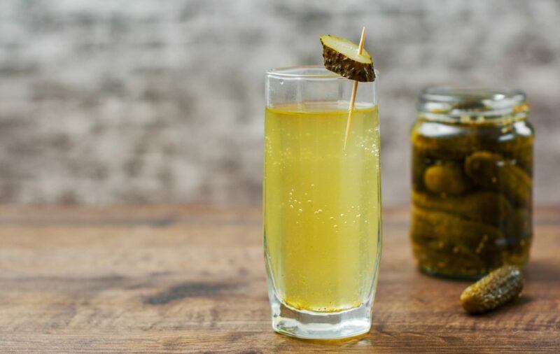 A glass of pickle juice garnished with a pickle, next to a jar of pickles