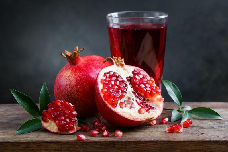 A glass of pomegranate juice on a table, next to a whole pomegranate, half a pomegranate, and a pomegranate piece