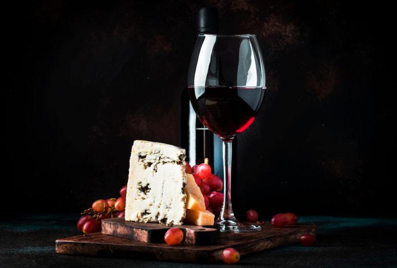 A glass and a bottle of port on a wooden board with cheese and fruit, against a black background