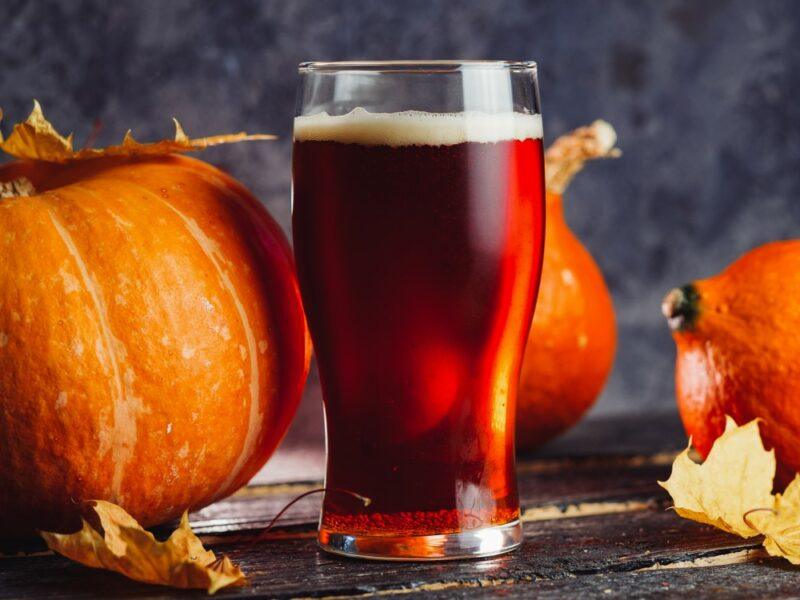 A glass of pumpkin beer on a table, next to some whole pumpkins