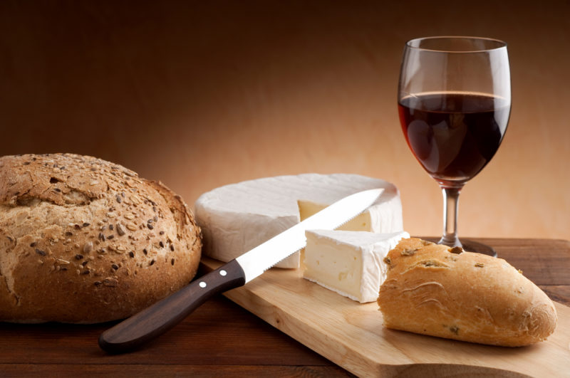 A wooden board with a glass of red wine, bread, and camembert cheese