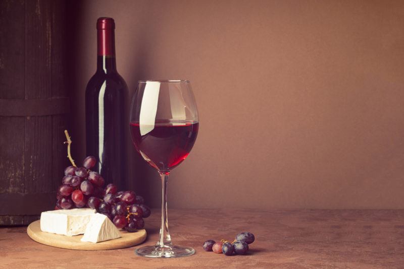 A glass of red wine on a wooden table, next to a bottle of the wine, grapes, and cheese