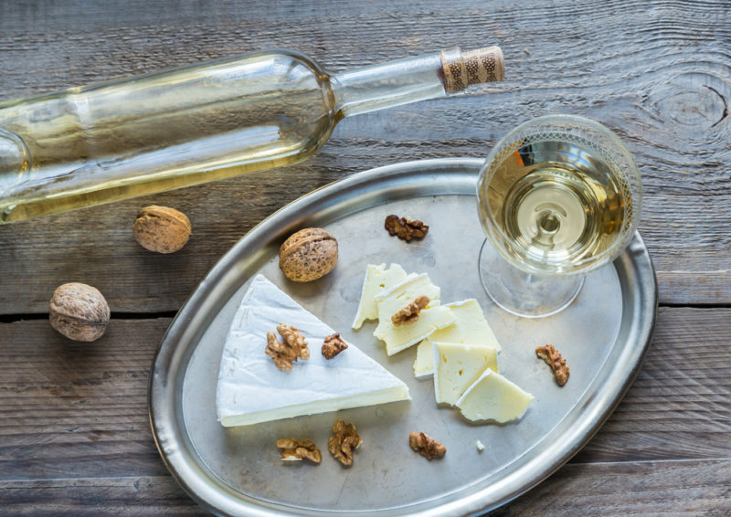 A metal tray with a wedge of camembert, some slices of cheese, nuts and a glass of riesling, next to a bottle of the wine on a table