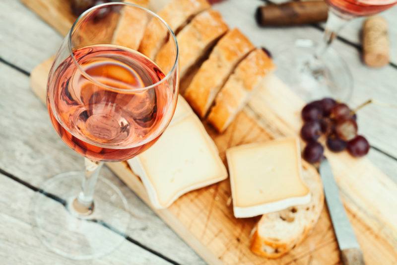 A glass of rose wine next to a cutting board with bread, cheese, grapes and a knife