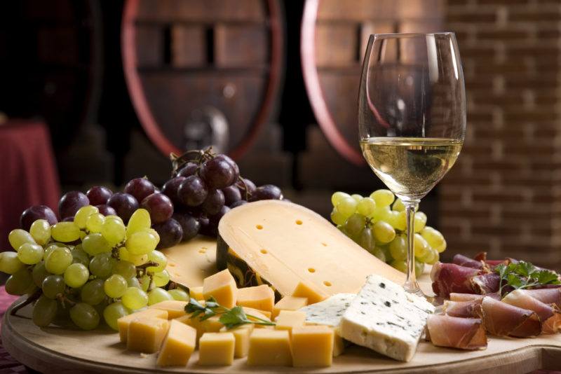 A glass of white wine on a wooden board, with various types of cheese, red grapes, and white grapes