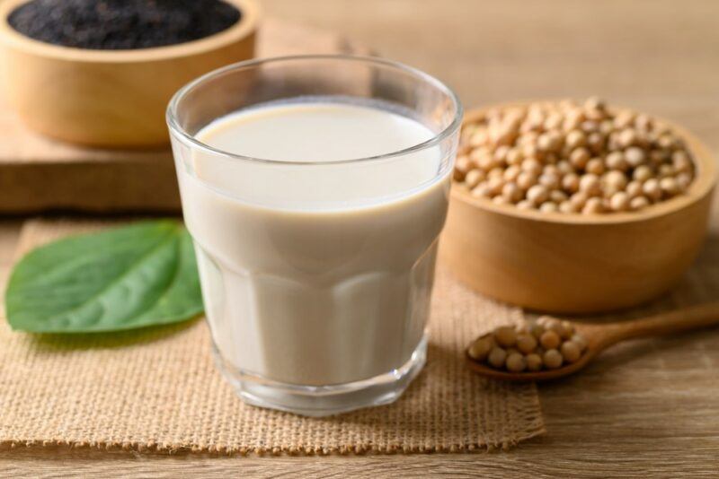 A glass of soy milk on a table, with a brown spoon and dish of soybeans in the background, plus a leaf and a bowl of black rice.