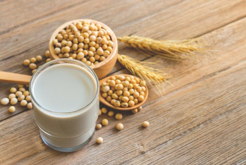 A glass of soy milk, next to a spoon of soybeans and a small brown container of soybeans