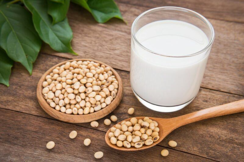 A wooden table with a glass of soy milk, a small flat wooden dish of soybeans, a wooden spoon containing soybeans, and some individual soybeans scattered on the table