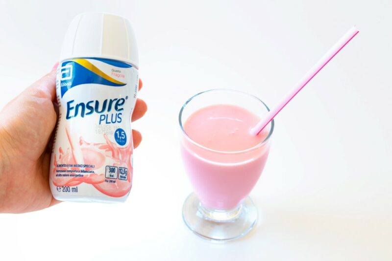 A hand holding a bottle of Ensure next to a glass filled with the pink shake