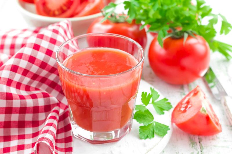 A wooden table that has a glass of tomato juice on top, along with a gingham placemat
