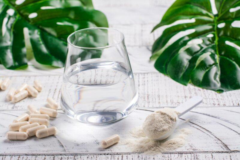 A glass of water, next to a scoop of collagen powder and some collagen supplements, with two large green leaves in the background