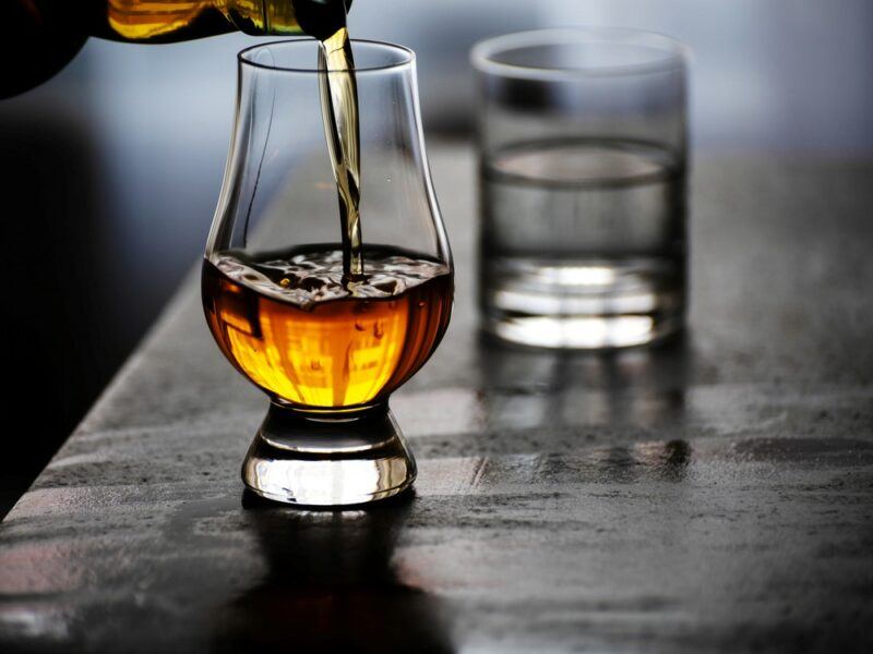 Someone pouring whiskey into a glass, with a glass of water in the background, implying that some of the water will be mixed into the whiskey