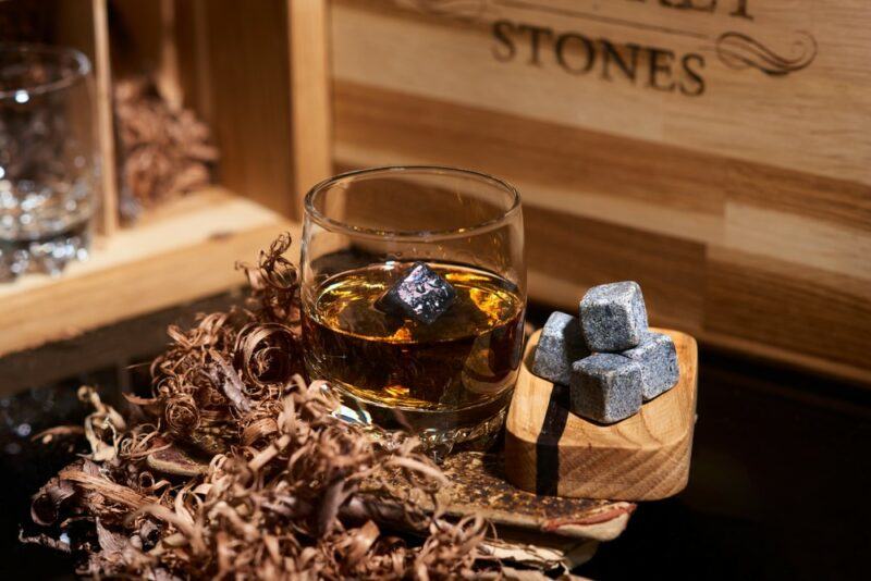 A glass containing whiskey and whiskey stones, in front of a wooden box and next to a small pile of more whiskey stones