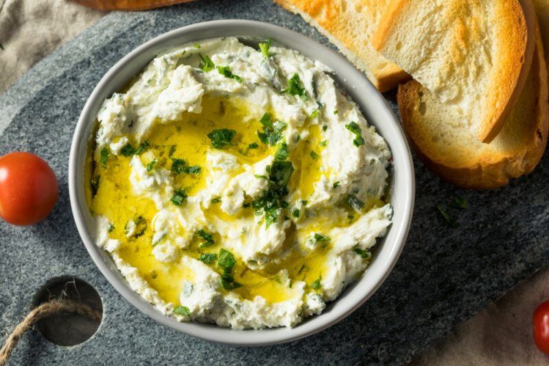 A bowl of goat cheese dip with oil and herbs on top, next to some bread