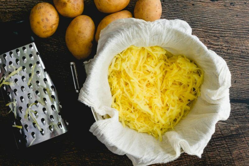 A grater next to some Yukon gold potatoes, next to a large bowl with cloth and grated potato.